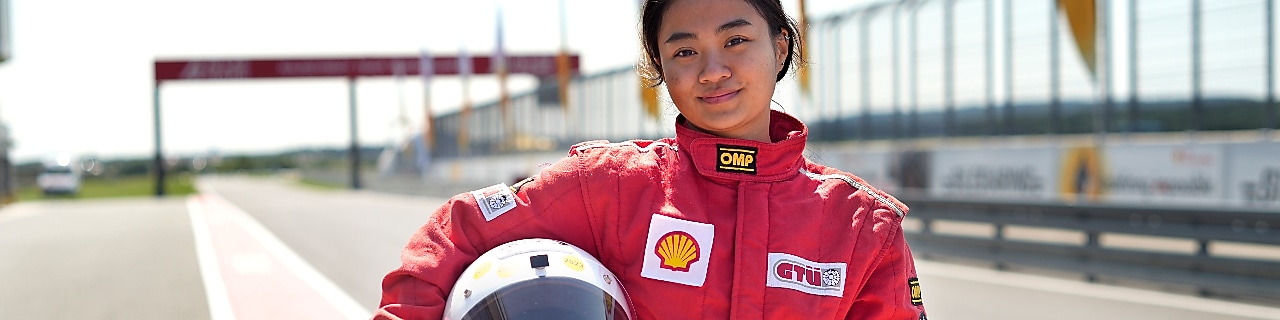 a young person wearing a driving suit and holding a race helmet posing in the pit lane of a race track