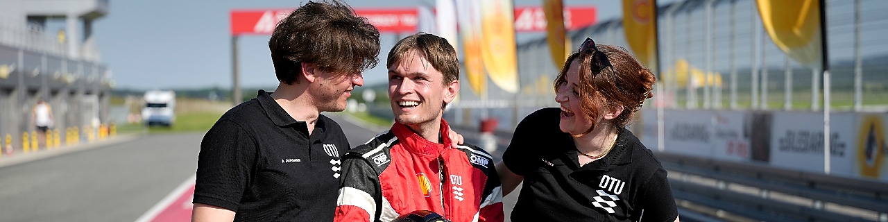 Three young people posing in the pit lane of a sunny race track, looking at each other, smiling.