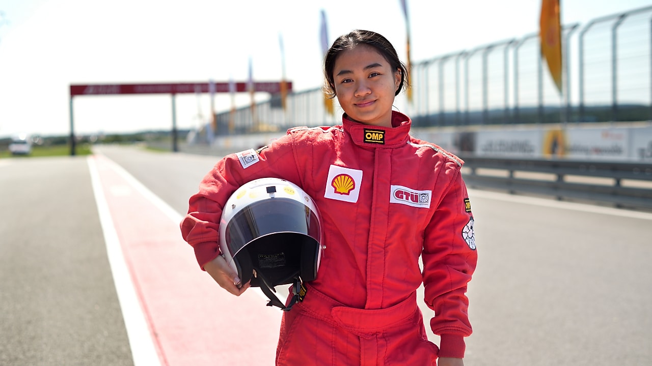 a young person wearing a driving suit and holding a race helmet posing in the pit lane of a race track