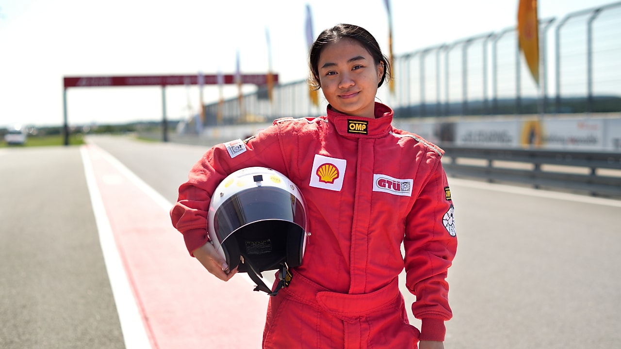 a young person wearing a driving suit and holding a race helmet posing in the pit lane of a race track