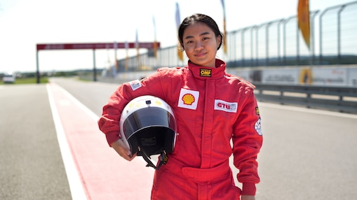 a young person wearing a driving suit and holding a race helmet posing in the pit lane of a race track