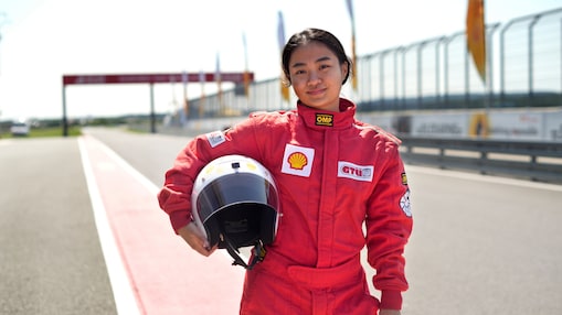 a young person wearing a driving suit and holding a race helmet posing in the pit lane of a race track.
