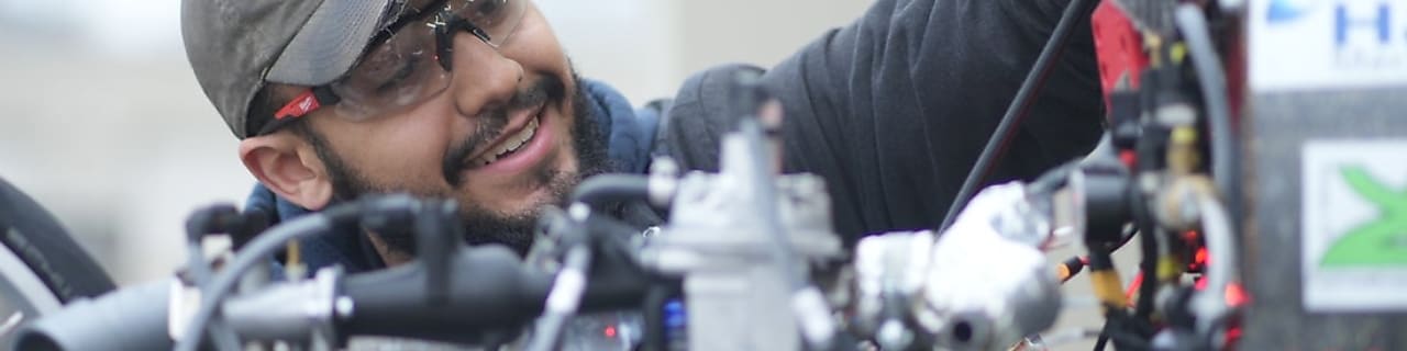 a young person wearing a cap, jacket and gloves working on a self-made car engine