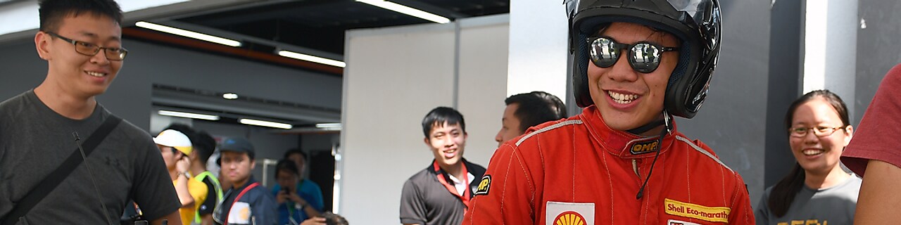 Candid photo of Shell Eco-marathon participant standing in his competition gear and smiling. He is surrounded by other Shell Eco-marathon participants who are also smiling and chatting.