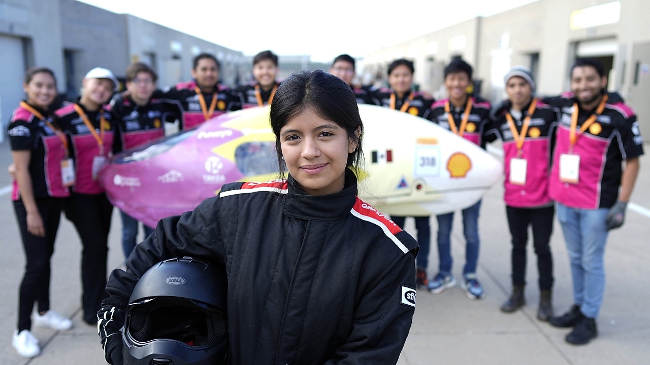 Person in driver’s suit standing in front of a group of people and a prototype vehicle.