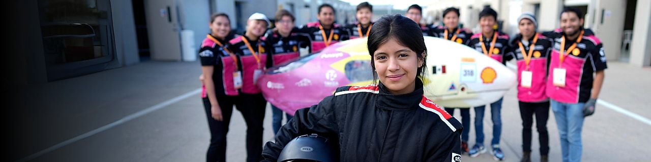 Person in driver’s suit standing in front of a group of people and a prototype vehicle.